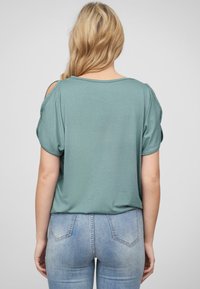 Haut vert à manches courtes avec une coupe décontractée, présentant des découpes aux épaules et une texture lisse, associé à un jean bleu clair.