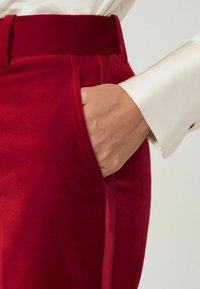 Červené zamatové nohavice s hladkou textúrou a bočnou kapsou, v kombinácii s bielou saténovou košeľou, ktorá zvýrazňuje pás a detaily šitia.