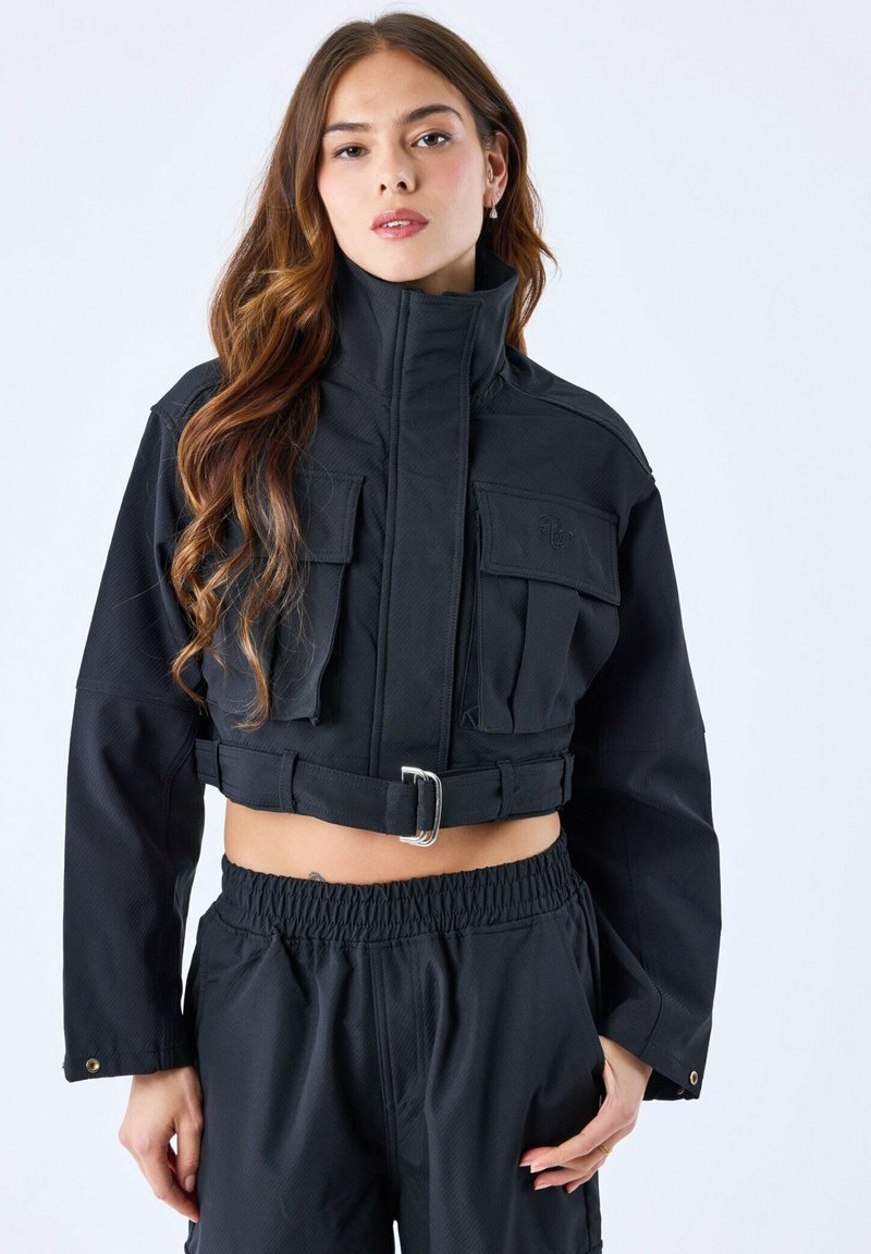 Zwarte cropped jacket met hoge kraag, twee voorzakken en riemdetail. De textuur lijkt gestructureerd met een matte afwerking.