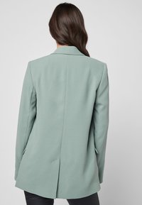 Donna con lunghi capelli scuri che indossa un blazer verde chiaro vista da dietro su uno sfondo grigio uniforme.