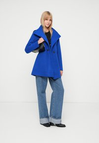 Cappotto blu doppiopetto con ampio colletto, dettagli con bottoni, abbinato a jeans a vita alta svasati e borsa nera. Texture liscia.