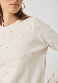 Svetle béžový pletený svetr s kulatým výstřihem, zdobený perlami a stříbrnými ozdobami, hladká textura a dlouhé rukávy.