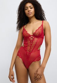 Ολόσωμο κορμάκι από κόκκινη δαντέλα με διαφανές σχέδιο και μοτίβα με κουκκίδες, κυματιστά άκρα, ανοίγματα και διασταυρούμενη λεπτομέρεια στο μπροστινό μέρος.