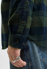 Hand mit einem großen, strukturierten Silberring, der ein grün-blau kariertes Hemd und blaue Jeans trägt.