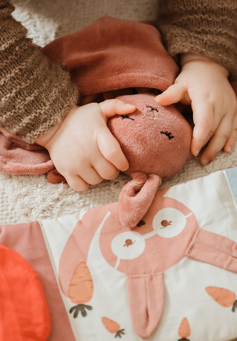 Jouet lapin doux et moelleux de couleur rouille tenu par de petites mains, posé sur une couverture texturée avec un tissu coloré et à motifs à proximité.