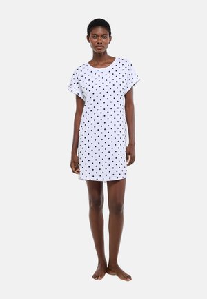 Femme debout, pieds nus, portant une robe blanche à manches courtes avec des pois noirs, sur un fond uni.