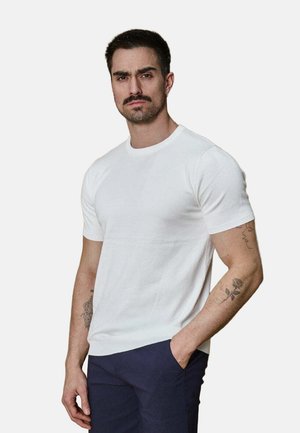 Homme avec moustache et tatouages portant une chemise blanche à manches courtes et un pantalon foncé, debout avec une main dans la poche sur un fond uni.