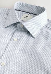 Light blue checked button-up shirt made of textured fabric, featuring a collar and white buttons, with a branded label inside the collar.
