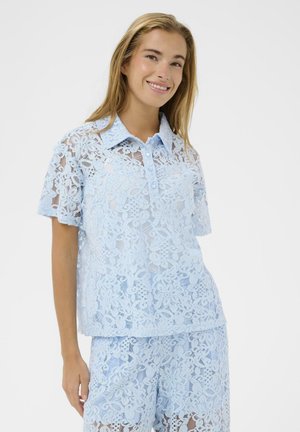 Šypsanti moteris, dėvinti šviesiai mėlyną, gėlių nėrinių trumpomis rankovėmis marškinius ir derančius šortus, stovinti priešais paprastą baltą foną.