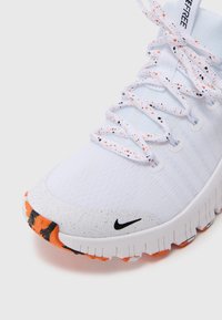 Sapatilha desportiva branca com sola e atacadores salpicados de laranja e preto, apresentando um pequeno swoosh preto da Nike perto da área do dedo do pé.