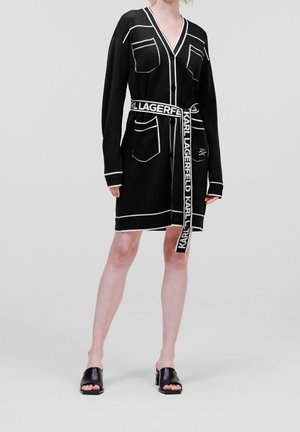 Personne portant une robe cardigan noire longueur genou avec bordure blanche, poches avant et ceinture de marque nouée à la taille, associée à des talons ouverts noirs.