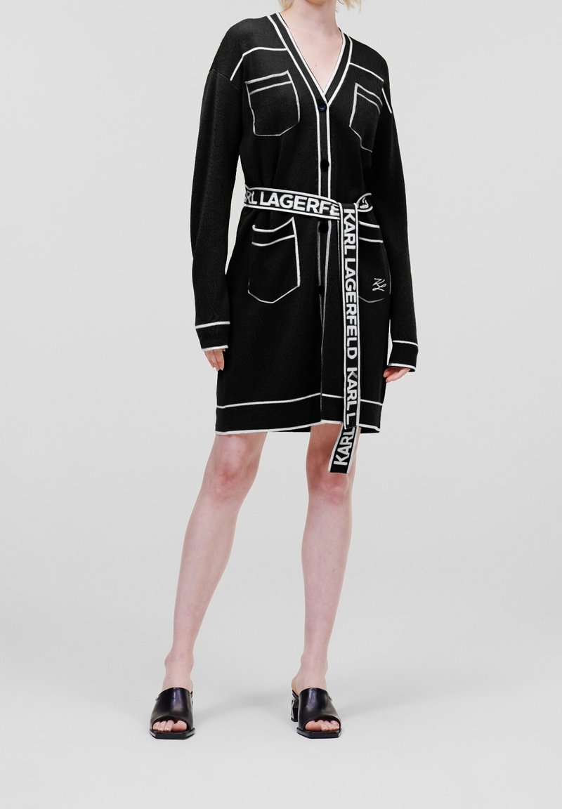 Personne portant une robe cardigan noire longueur genou avec bordure blanche, poches avant et ceinture de marque nouée à la taille, associée à des talons ouverts noirs.