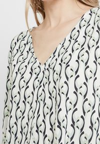 Blouse blanche à col en V, avec un motif abstrait noir et vert pâle. Tissu léger et texturé avec un tombé doux.