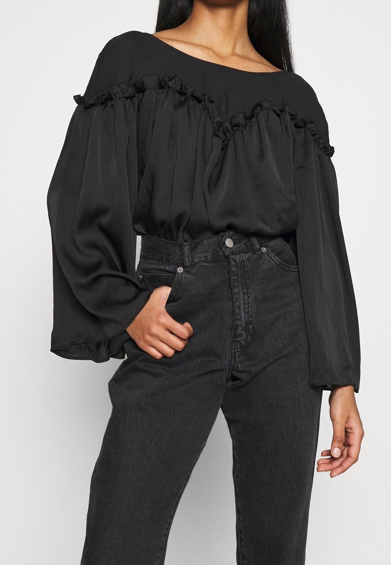 Mujer vestida con una blusa negra de hombros al aire con detalles de volantes y mangas globo, combinada con unos vaqueros de mezclilla negros de cintura alta.