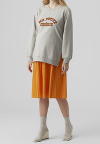 Grå sweatshirt med texten "MA PETITE CHÉRIE", kombinerad med en plisserad orange kjol. Modellen bär grå revärstrumpor och kilklackar.