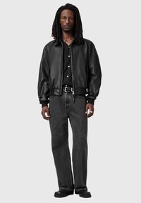 Czarna skórzana kurtka bomber o luźnym kroju, czarna koszula na guziki, luźne ciemne dżinsy i czarne buty. Gładka faktura i minimalistyczne okucia.