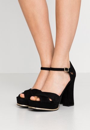 Czarne zamszowe sandały na wysokim obcasie z węzłowym wzorem na górze, z paskiem na kostce i masywnym obcasem. Charakteryzują się złotym akcentem na krawędzi podeszwy.