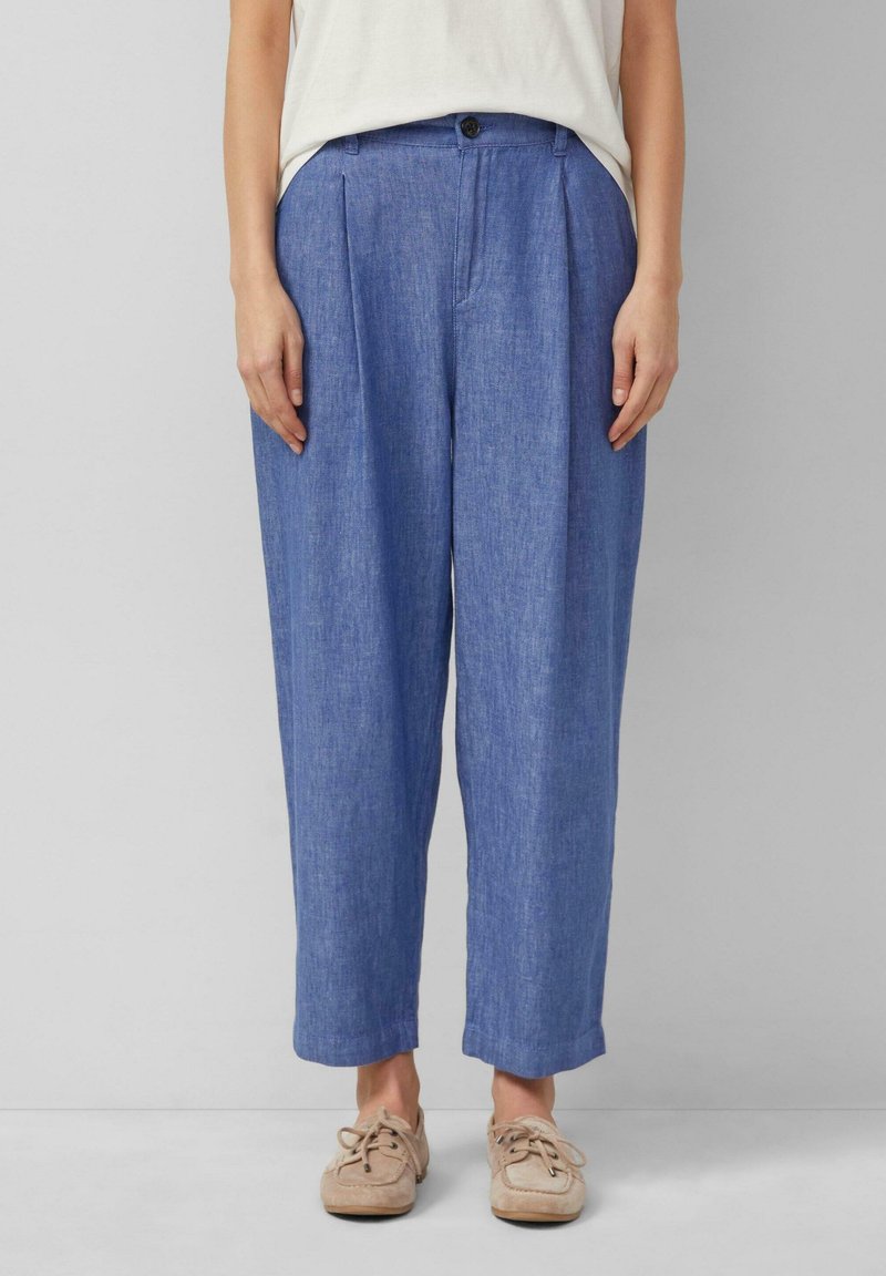 Femme portant un pantalon bleu ample, des chaussures beiges à lacets et un haut blanc sur un fond uni.