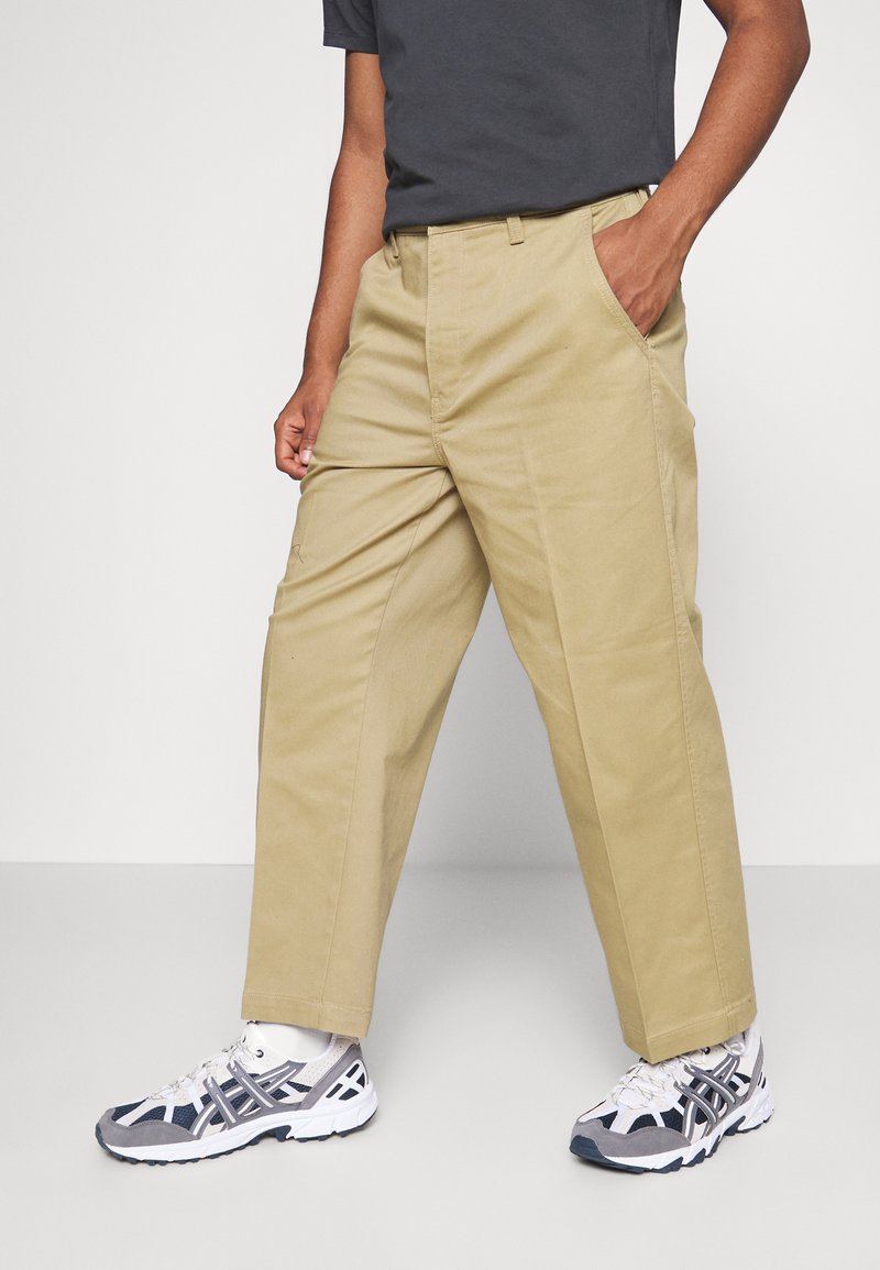 Pantaloni cargo beige con tasche laterali, taglio dritto e tessuto misto cotone, abbinati a scarpe da ginnastica grigie e bianche.