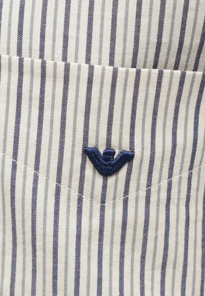 Stripet stoff i hvit og marineblå med brodert vinge-logo. Teksturen er jevn, og designet har vertikale linjer og en lomme.