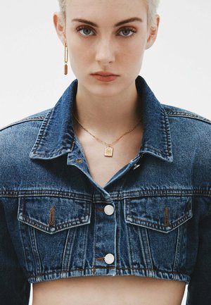 Chaqueta de mezclilla recortada en azul medio con botones plateados, dos bolsillos en el pecho y un cuello. Collar de oro con un pendant cuadrado.