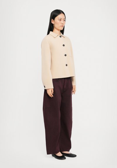 Chaqueta corta beige con botones negros, un bolsillo; combinada con pantalones sueltos de un burdeos oscuro y zapatos planos negros.