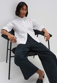 Camicia bianca a colletto con maniche corte, abbinata a pantaloni a gamba larga blu navy, seduta su una sedia nera contro uno sfondo chiaro.