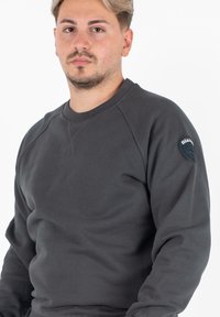 Szary sweter z okrągłym dekoltem, rękawami raglanowymi i teksturowanym wykończeniem. Posiada naszywkę z logo na lewym ramieniu.
