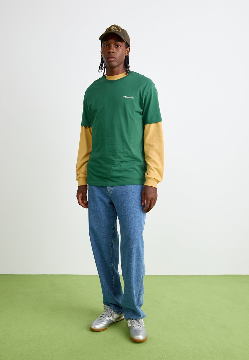 T-shirt verde a maniche corte indossato sopra una maglietta gialla a maniche lunghe, abbinato a jeans blu e scarpe da ginnastica argentate. Berretto marrone con dettaglio del logo.