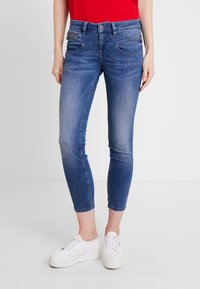 Jeans skinny de denim azul con una textura suave, que cuentan con bolsillos delanteros, detalles de costura y una cintura de altura media, combinados con zapatillas blancas.