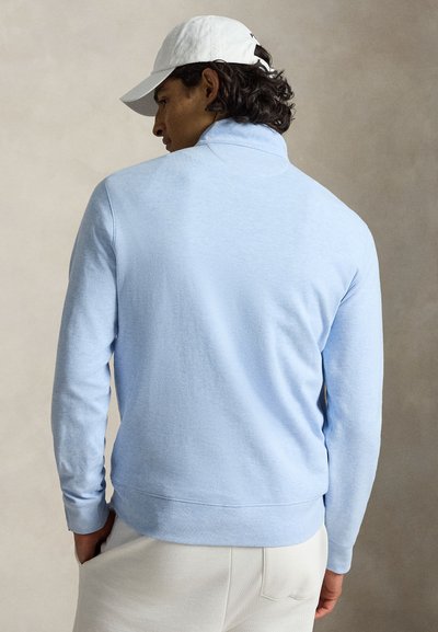 Moški s temnimi kodrastimi lasmi, ki nosi belo kapo, svetlomodro srajco z dolgimi rokavi in krem hlače, prikazan od zadaj na nevtralnem ozadju.