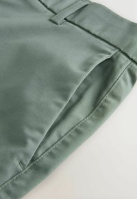 Detailný záber na svetlozelené nohavice z tkaniny ukazujúce predný vrecko, švy a detail očiek na opasok na svetlom pozadí.