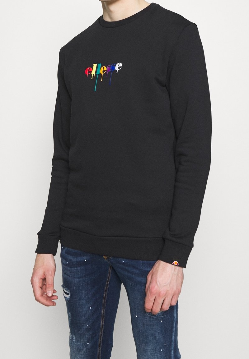 Felpa nera con polsini e fondo a coste, caratterizzata da un logo multicolore che gocciola sul davanti. Indossata con jeans blu strappati.