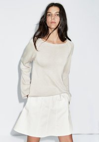 Beżowy sweter dzianinowy z szerokim dekoltem i długimi rękawami, zestawiony z białą plisowaną spódnicą w kształcie litery A. Gładka struktura, swobodny styl.