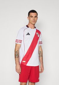 Vit fotbollströja med ett diagonalt röd streck, med svarta detaljer och Adidas-logotypen. Kombineras med röda shorts.