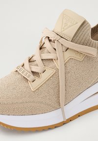 Beige knit sneaker with thick white midsole, gum rubber outsole, beige laces, and Guess logo on tongue and lace tag.