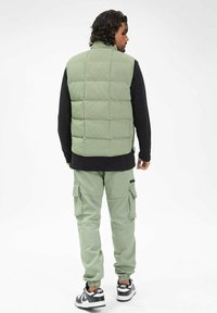 Veste rembourrée vert clair avec un motif en carré, portée sur une chemise à manches longues noire, associée à un pantalon cargo assorti et des baskets.