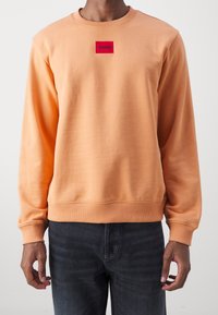 Pomarańczowa bluza z okrągłym dekoltem, ściągaczami na rękawach i dole. Na piersi znajduje się czerwony prostokątny naszywka z logo "HUGO". Gładka faktura.