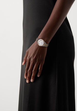 Persoon draagt een zilveren en gouden polshorloge met een witte wijzerplaat en Romeinse cijfers, gekleed in een zwarte mouwloze outfit.