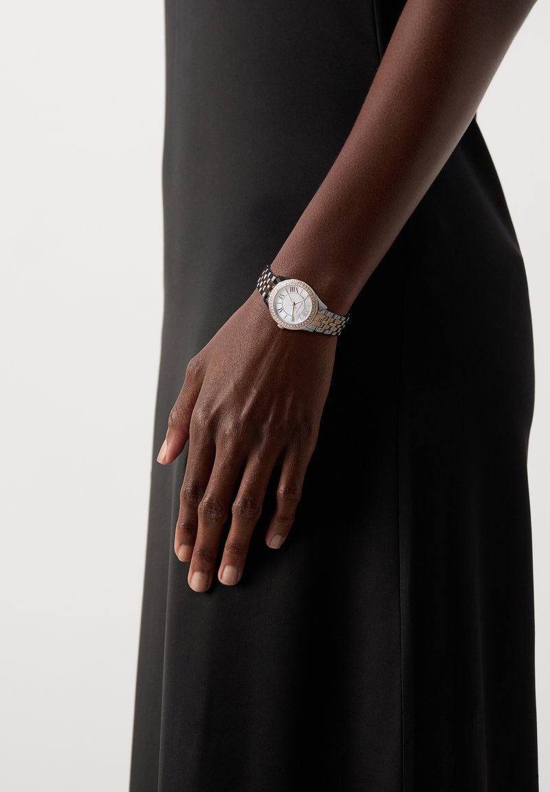 Person trägt eine silberne und goldene Armbanduhr mit weißem Zifferblatt und römischen Zahlen, bekleidet mit einem schwarzen ärmellosen Outfit.