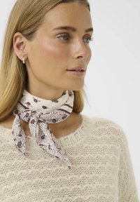 Écharpe légère crème avec des motifs floraux et rayés, nouée autour du cou. Portée avec un pull en tricot beige texturé.