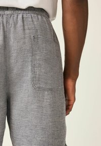 Nahaufnahme einer Person, die graue, strukturierte Hosen mit einem elastischen Bund und einer quadratischen Gesäßtasche trägt und mit einem sichtbaren Arm steht.