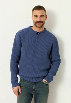 Mann mit Schnurrbart, der einen blauen Strickpullover und Jeans trägt, steht mit einer Hand in der Tasche vor einer weißen Wand. Sichtbare Handtätowierungen sind zu erkennen.