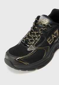Svart atletisk sneakersko med mesh- og syntetiske materialer. Gullfargede detaljer og snøringer forbedrer designet. Bred, teksturert såle for støtte.