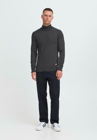 Uomo in piedi, che indossa un maglione grigio scuro a collo alto, pantaloni neri e scarpe bianche con strisce nere, su sfondo bianco.