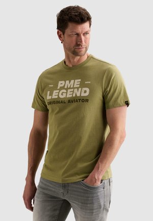 Άνδρας με ελιά πράσινο μπλουζάκι "PME Legend Original Aviator", γκρι τζιν, κοιτάει δεξιά με το ένα χέρι στην τσέπη σε λευκό φόντο.