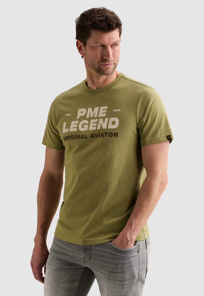 Άνδρας με ελιά πράσινο μπλουζάκι "PME Legend Original Aviator", γκρι τζιν, κοιτάει δεξιά με το ένα χέρι στην τσέπη σε λευκό φόντο.