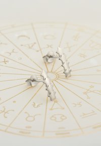 Silverstjärnformade örhängen med en polerad finish, som har sex länkade stjärnor i en postdesign, mot en guldkantad astrologisk bakgrund.
