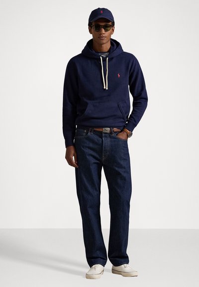 Sudadera navy con cordón, logo en el pecho, combinada con jeans oscuros y zapatillas blancas, accessorios con gafas de sol y una gorra.