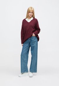 Pull oversize bordeaux avec un profond décolleté en V, associé à un jean bleu clair à jambes larges et des baskets blanches sur un fond uni.